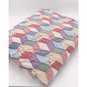 May include: A folded quilt featuring a geometric tumbling block pattern composed of various fabric patches. The design includes shades of pink, purple, blue, and cream, with some patches displaying small floral prints and textured fabric patterns.