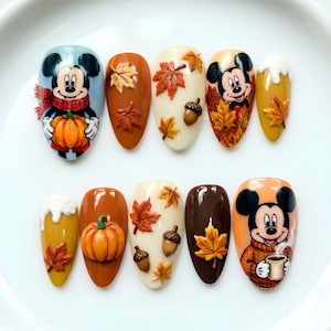 May include: A set of ten press-on nails featuring autumn themes, including Mickey Mouse illustrations, orange pumpkins, brown acorns, and various maple leaves in shades of red, yellow, and brown, set against cream, brown, and mustard yellow backgrounds.