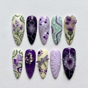 May include: A set of ten almond-shaped press-on nails featuring various designs in shades of purple, lavender, cream, and sage green. Embellishments include 3D crystals, a silver dragonfly charm, raised floral patterns, and painted motifs.