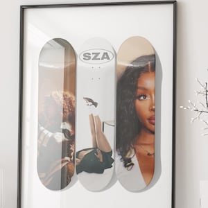 May include: A framed wall art print featuring three vertical skateboard decks arranged side-by-side. The centre deck is white with the text SZA and a small graphic, while the outer decks display photographic portraits of a person with long dark hair.