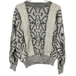 May include: A long-sleeve knit sweater featuring a cream and charcoal gray geometric pattern. The garment includes vertical cable knit panels on the front, a ribbed crew neckline, and ribbed cuffs and hem in a speckled gray knit texture.