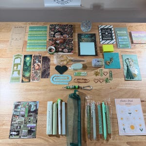 May include: A collection of stationery items including a squirrel-themed notebook, various bookmarks, pens, a green mesh pencil case, sticker sheets, paper clips, and a book review card. The items are arranged on a wooden surface with a logo sign above.