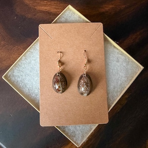 May include: A pair of dangle earrings featuring small, oval-shaped brown cowrie shells with white speckled patterns. The shells are attached to gold-toned metal fishhook ear wires and are displayed on a rectangular brown cardboard jewelry card.