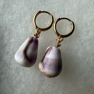 May include: A pair of gold hoop earrings featuring dangling natural cowrie shells. Each shell displays a smooth, polished surface with a gradient of white and deep purple hues. The shells are securely attached to the gold hoops with small wire loops.