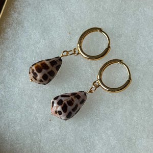 Puede incluir: Un par de pendientes de aro dorados, cada uno con un colgante de concha en forma de lágrima. Las conchas son de color beige claro con un patrón distintivo de color marrón oscuro en forma de cuadrícula en sus superficies. Los aros son de metal pulido.