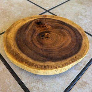 May include: A circular wooden slab featuring a dark brown center with visible growth rings and a lighter, yellowish-tan outer edge. The surface displays a prominent natural knot near the top, showcasing the raw texture and grain of the wood.