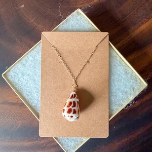 May include: A gold-toned metal chain necklace featuring a teardrop-shaped seashell pendant. The shell is white with a pattern of reddish-brown spots. The necklace is displayed on a rectangular brown card with two small holes for the chain.
