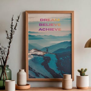May include: A framed vertical wall art print featuring a mountain landscape with a person standing on a peak. The text DREAM BELIEVE ACHIEVE is printed in a gradient color scheme at the top. The frame is light wood, and the image shows blue mountains.