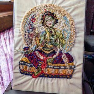 Puede incluir: Una pieza rectangular de tela color hueso que presenta un bordado detallado y multicolor de una figura sentada. La figura está rodeada por un halo circular y se asienta sobre una base estampada, en tonos verde, azul, rojo y dorado.
