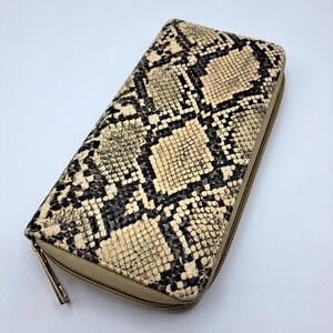 Puede incluir: Una cartera rectangular con cremallera que presenta un patrón texturizado de piel de serpiente en tonos crema, beige y negro. La cartera tiene un tirador de cremallera de metal dorado unido a una cinta de tela beige a juego en el borde.