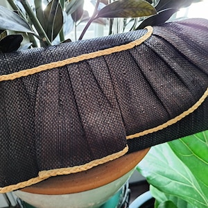 Könnte beinhalten: Eine dunkelbraune, geflochtene Clutch mit einer plissierten Frontklappe und einem hellbeigen, geflochtenen Randbesatz. Das Material hat eine texturierte, strohähnliche Optik mit einer strukturierten, rechteckigen Form und einem geschwungenen Klappenverschluss.