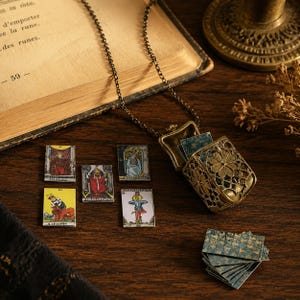 Pode incluir: Um pingente de relicário de metal em tom de bronze numa corrente, com um design intrincado de filigrana floral. O relicário aberto contém cartas de tarot em miniatura, com cartas de tarot pequenas adicionais exibidas perto com figuras.