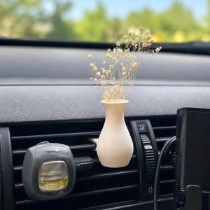 May include: A small, matte off-white vase attached to a black car air vent, containing a cluster of delicate, dried cream-colored baby's breath flowers. A gray car air freshener is clipped to the vent slats to the left of the vase.