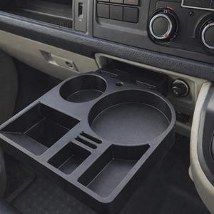 Peut inclure: Un porte-gobelet et plateau de rangement en plastique noir pour véhicule, déployé depuis la console du tableau de bord. Le plateau comprend un grand porte-gobelet circulaire, un plus petit et trois compartiments de rangement rectangulaires.
