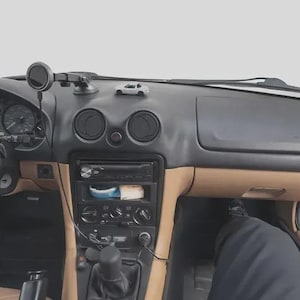 May include: The interior of a vehicle featuring a black dashboard with tan lower panels, a central console with a radio and climate controls, a black steering wheel, a gear shift lever, and a small white toy car model placed on the dashboard top.