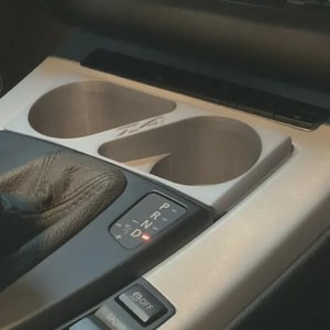 May include: A light gray dual cup holder insert installed in a vehicle center console. The insert features two circular openings and is positioned next to a black gear shifter and a gear selection panel displaying P, R, N, D, and M/S settings.