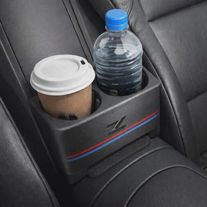 Puede incluir: Un portavasos doble negro con textura de metal cepillado, un logotipo Z prominente y franjas decorativas horizontales rojas y azules. La unidad sostiene un vaso de café de papel marrón con tapa blanca y una botella de agua de plástico transparente.