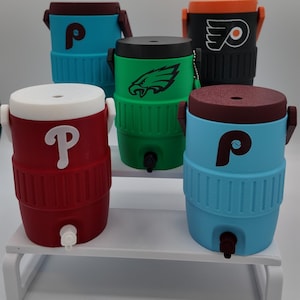 May include: Five miniature beverage coolers with spigots and lids, featuring various sports team logos including the Philadelphia Phillies P, Philadelphia Eagles eagle, and Philadelphia Flyers winged P, in red, blue, green, and black colors.