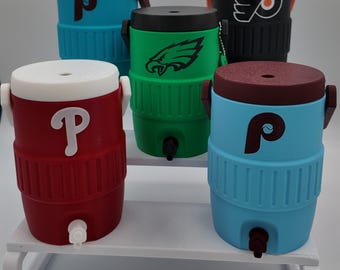 May include: Five miniature beverage coolers with spigots and lids, featuring various sports team logos including the Philadelphia Phillies P, Philadelphia Eagles eagle, and Philadelphia Flyers winged P, in red, blue, green, and black colors.