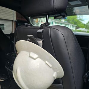 May include: A white hard hat suspended from a black plastic hook attached to the metal headrest post of a black leather vehicle seat. The hard hat features a smooth, rounded shell with a brim and molded structural ridges on the exterior surface.