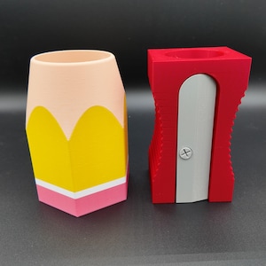 Peut inclure: Un organisateur de bureau en forme de crayon avec un sommet pêche, un milieu jaune, une bande blanche et une base rose, placé à côté d'un récipient rouge en forme de taille-crayon avec une lame argentée et une vis centrale visible.
