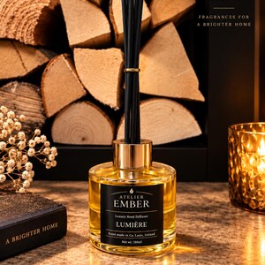 Może przedstawiać: Szklany dyfuzor zapachowy ze złotym kołnierzem i czarnymi patyczkami. Czarna etykieta zawiera napis Atelier Ember, Luxury Reed Diffuser, Lumière, Hand made in Co. Laois, Ireland, Net wt. 100ml. Butelka zawiera złoty płyn zapachowy.