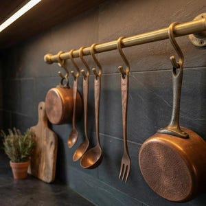 May include: A brass-toned metal rail mounted horizontally with six matching S-shaped hooks. Hanging from the hooks are a small copper pot, a copper spoon, a copper ladle, a copper fork, and a larger copper pot with a long handle.