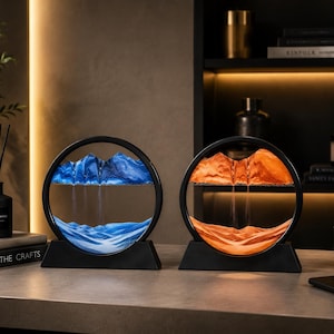 Pode incluir: Dois expositores circulares de arte em areia com molduras e bases pretas. Um apresenta areia azul formando montanhas e vales, enquanto o outro contém areia laranja criando paisagens em camadas semelhantes. Ambos estão sobre uma superfície plana.