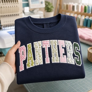 May include: A navy blue crewneck sweatshirt featuring the word PANTHERS in large, arched, fabric-appliqued letters. Each letter is crafted from a different patterned fabric, including pink polka dots, green floral, and pink and white stripes.