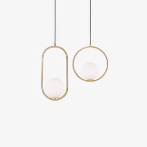 May include: Two pendant lights suspended by thin black cords. One fixture features a vertical gold-toned oval frame holding a white spherical glass globe, while the other consists of a circular gold-toned frame with a matching white glass globe.