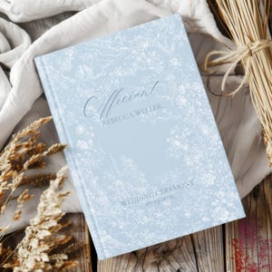 May include: A light blue hardcover book with a white toile de jouy floral pattern. The cover features the text Officiant, REBECCA WELLER, and WEDDING CEREMONY 07-15-2026 in a dark blue script and serif font, centered on the front cover.