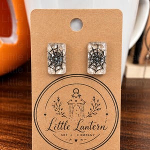 Può includere: Orecchini a lobo rettangolari con un disegno di ragnatela nera incastonato in resina trasparente con glitter argentati. Gli orecchini sono esposti su un cartoncino marrone con la scritta Little Lantern Art + Company.