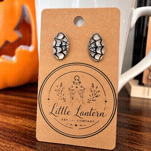 May include: A pair of oval stud earrings featuring a black spiderweb pattern on a clear, glittery background. The earrings are attached to a brown cardboard display card printed with the text Little Lantern Art Company.