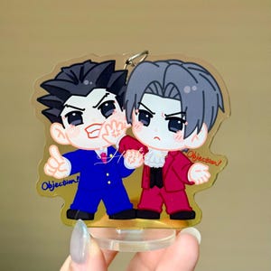 May include: A clear acrylic standee featuring two chibi-style characters. One character has spiky black hair and a blue suit, the other has grey hair and a red suit. Both figures have the word Objection! printed in blue and red text respectively.
