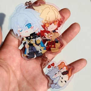 May include: A clear acrylic keychain featuring two chibi-style anime characters with spiky hair, one with light blue hair and the other with blonde hair, attached to a smaller secondary charm depicting three small cartoon animals hugging.