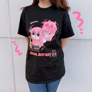 May include: A black short-sleeve t-shirt featuring a pink graphic of an anime character holding a guitar with the text "BOCCHI" and "SOCIAL BATTERY 0%" printed in pink. The design includes a battery icon and pink Japanese characters.