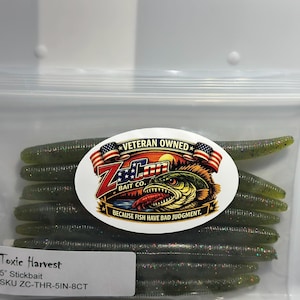 May include: A clear plastic resealable bag containing eight olive green, glitter-flecked 5-inch stickbait fishing lures. A white oval label features the Z-Con Bait Co. logo, a fish illustration, and the text: Veteran Owned, Because Fish Have Bad Judgment.