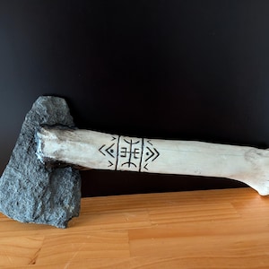 May include: A stone axe featuring a dark gray, rough-textured stone head securely attached to a light-colored, bone-like handle. The handle is decorated with black, hand-drawn geometric symbols and linear patterns centered along its length.