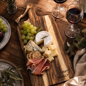 May include: A rectangular wooden serving board with a handle and a jute rope loop, featuring the engraved text "Williams FAMILY". The board is arranged with green grapes, a round soft cheese, blue cheese, crackers, and prosciutto slices.