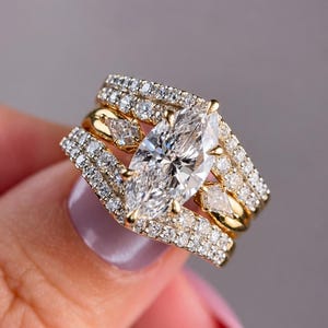 May include: A gold ring set featuring a large marquise-cut center stone flanked by two smaller marquise-cut stones. The bands are encrusted with multiple rows of small, clear, round-cut stones, creating a layered, multi-band appearance.