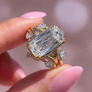 May include: A gold ring featuring a large rectangular emerald-cut center stone surrounded by a halo of smaller round brilliant-cut diamonds. The band is crafted from polished yellow gold and includes additional diamond accents along the setting.