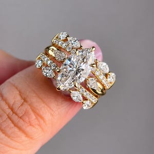 May include: A gold ring set featuring a large marquise-cut center stone surrounded by smaller marquise and round-cut stones. The design includes multiple bands with pavé-set diamonds, creating a layered appearance with a polished gold finish.