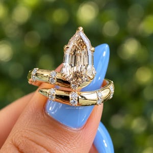 May include: A gold bypass ring featuring a large pear-shaped champagne-coloured centre stone and a band accented with small round white diamonds. The ring is engraved with the text CRYSTLLE MILANO on the inner surface of the gold band.