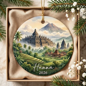 May include: A round ceramic ornament featuring a watercolor-style illustration of the Borobudur temple complex, lush green rice terraces, and a large mountain in the background. The name Hanna and the year 2026 are printed in white script at the bottom.