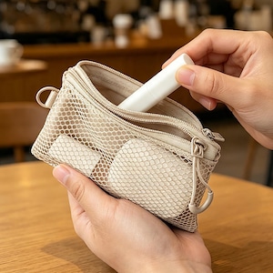 May include: A beige mesh zippered pouch with two compartments, featuring a fine grid pattern and matching beige fabric trim. The pouch is held open to reveal a smooth interior, and a small white cylindrical object is being placed inside.