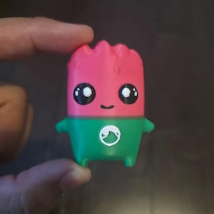 May include: A small, upright figurine shaped like a watermelon slice with a pink top and a green bottom. It features two large black eyes with white highlights, a simple black smile, and a small white circular logo on the green section.