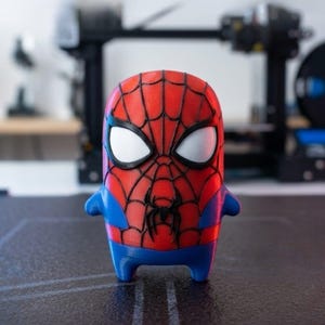 May include: A small, stylised figurine of a superhero character with a red and blue colour scheme. The figure features a prominent black web pattern across the red head and chest, large white eye lenses with black borders, and a small black spider emblem.