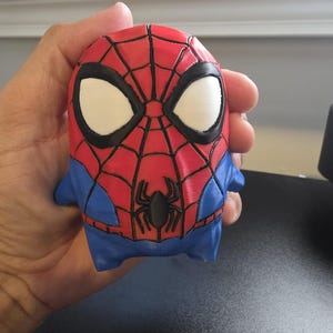 Könnte beinhalten: Eine kleine, stilisierte Figur eines Superhelden mit einem roten und blauen Farbschema. Der Kopf zeigt eine rote Maske mit schwarzem Netzmuster, großen weißen Augen und einem kleinen schwarzen Spinnenemblem auf der Brust. Der Körper ist rund.