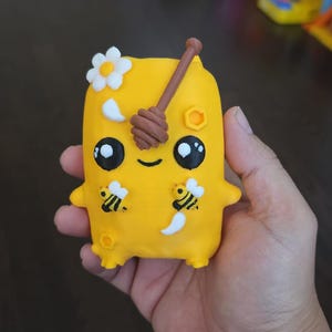 Könnte beinhalten: Eine gelbe, rechteckige, tonartige Figur mit einem lächelnden Gesicht, großen schwarzen Augen und zwei kleinen Armen. Sie ist mit einer weißen Blume, einem braunen Honiglöffel, zwei kleinen Bienen und Wabenmustern auf der glatten Oberfläche verziert.
