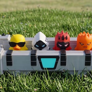 May include: A light gray rectangular storage crate with black accents and a central blue panel containing four small stylized figures: a yellow duck with sunglasses, a white hooded character, a red horned figure, and an orange creature.